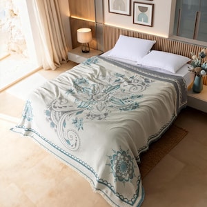 May include: A cream-colored bedspread featuring a large teal and gray floral mandala pattern in the center, with smaller matching mandala motifs at the corners and a decorative border. The fabric has a textured finish and covers a bed with two white pillows.