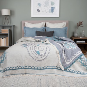 May include: A bed featuring a cream-colored throw blanket with a large blue paisley pattern and a scalloped border. The bed is styled with two light blue pillows, two white pillows, and a small dark gray rectangular pillow, all with fringe.