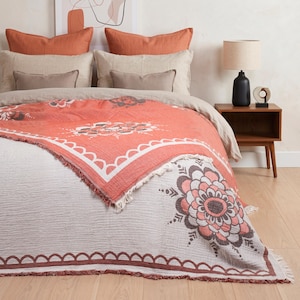 May include: A textured throw blanket featuring a coral and white floral mandala pattern with a fringed edge. The blanket is draped over a bed with beige and coral pillows, positioned next to a wooden nightstand with a black lamp and a small sculpture.