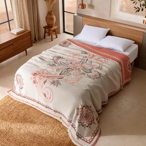 May include: A cream-colored bedspread featuring a large, intricate floral mandala pattern in shades of terracotta and dark gray. The textile has a textured surface, a wide terracotta border, and a fringed edge draped over a white mattress.