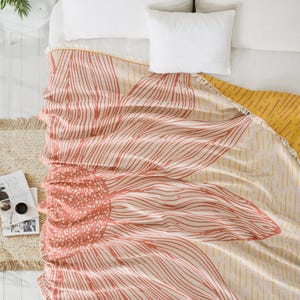 Puede incluir: Una manta rectangular con un gran diseño floral estilizado en color rosa coral con detalles de líneas blancas sobre un fondo crema. La manta tiene un reverso de color amarillo mostaza con un patrón de rayas y un borde con flecos arriba.