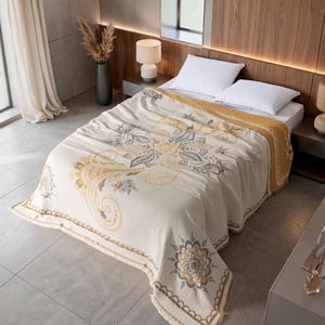 May include: A cream-colored bedspread featuring a large, central mandala-inspired pattern in shades of mustard yellow and gray. The textile has a textured surface, a solid mustard yellow border, and a fringed edge, draped over a white bed.