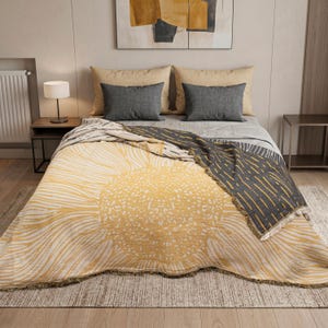 May include: A reversible bed throw featuring a large yellow sunflower graphic on one side and a dark grey background with yellow dashed lines on the other. The throw has a fringed edge and is draped over a bed with two beige and two grey pillows.