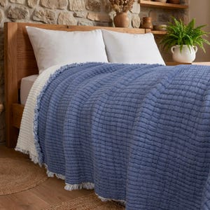 May include: A reversible bed throw featuring a textured blue side and a solid white side. The blanket has a frayed, fringed edge detail along the perimeter and is draped over a wooden bed frame with white pillows.