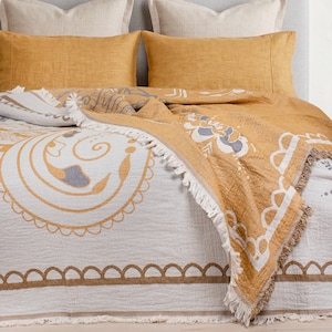May include: A textured throw blanket featuring a mustard yellow and white color palette with a large circular floral pattern and a scalloped border. The blanket has frayed fringe edges and is draped over a bed with two mustard yellow pillows.