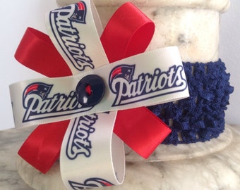 May include: A white ribbon with the word "Patriots" printed on it in blue and red. The ribbon is tied into a bow with a red ribbon. The bow is sitting on a white surface with a blue crocheted fabric.