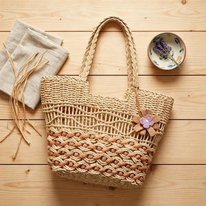 Könnte beinhalten: Eine beige geflochtene Tragetasche mit einem komplizierten Makramee-Muster und geflochtenen Griffen. Die Tasche enthält einen dekorativen Blumenanhänger mit einem violetten Zentrum an der Seite. Das Design kombiniert hellbraune und dunkelbraune Kordeln.