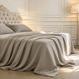 Könnte beinhalten: Eine beige Tagesdecke, die über ein Bett mit gepolstertem Kopfteil und zwei passenden Kissen drapiert ist. Die Bettwäsche hat eine strukturierte Stoffoberfläche, die über die Seiten der Matratze fällt und weiche Falten im warmen Licht bildet.
