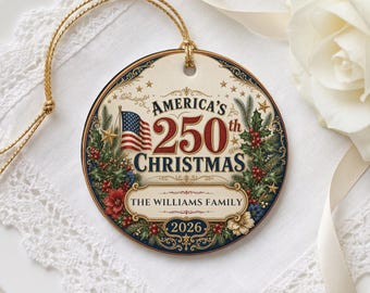 May include: A round ceramic ornament featuring a gold string, decorated with a red, white, and blue American flag, holly leaves, and berries. The text reads America's 250th Christmas, The Williams Family, and 2026 in a decorative vintage style.