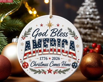 May include: A round white ceramic ornament hanging by a string. The text reads God Bless AMERICA 1776 2026 Christmas Comes Home. The word AMERICA is filled with a red, white, and blue star-spangled banner pattern. Decorated with red berries.