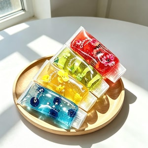 Peut inclure: Quatre tubes souples transparents étiquetés Chaboky, remplis de gel de différentes couleurs contenant des éléments floraux incrustés et des petites étoiles. Les tubes rouges, verts, jaunes et bleus sont disposés sur un plateau en bois.