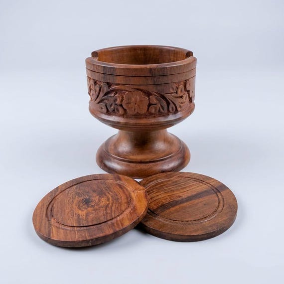 Hand-carved solid wood coasters on a goblet-style pedestal stand — the carved floral detail makes this feel like a craft object, not a commodity.