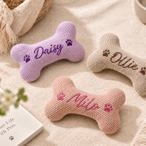 May include: Three bone-shaped pet toys made of ribbed corduroy fabric in lavender, beige, and dusty pink. Each toy features embroidered names: Daisy in purple, Ollie in brown, and Milo in pink, accompanied by small embroidered paw prints.