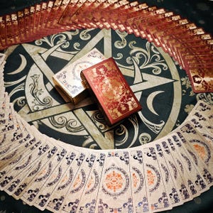 Pode incluir: Um baralho de cartas disposto em leque circular sobre uma superfície escura com um grande design de pentagrama. Duas caixas de cartas retangulares estão colocadas no centro, uma com um padrão floral vermelho escuro e dourado e a outra creme.