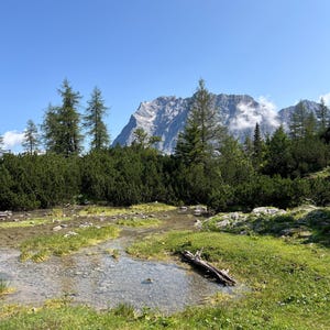 Könnte beinhalten: Eine Landschaft mit einem flachen, klaren Bach, der durch eine grasbewachsene Wiese mit verstreuten Steinen und einem umgefallenen Baumstamm fließt. Im Hintergrund erhebt sich ein großer, schroffer Berggipfel vor einem blauen Himmel.