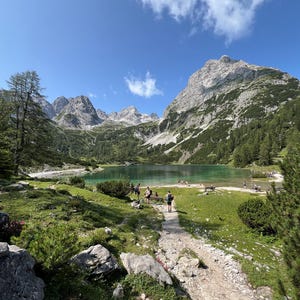 Könnte beinhalten: Eine malerische Landschaft mit einem türkisfarbenen Alpensee, umgeben von zerklüfteten, felsigen Berggipfeln unter einem klaren blauen Himmel. Ein Kieswanderweg führt zum Wasser, gesäumt von grünen Wiesen, Kiefern und verstreuten grauen Felsen.