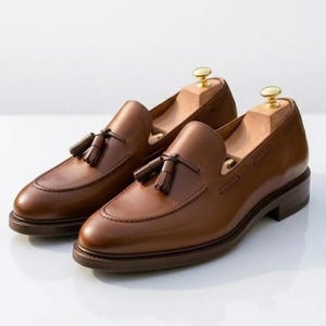 May include: A pair of brown leather tassel loafers featuring a rounded toe, a stitched apron front, and a low stacked heel. Each shoe contains a light-colored wooden shoe tree with a gold-toned metal knob at the heel for maintaining shape.