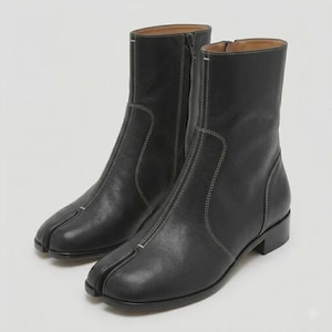 May include: A pair of black leather ankle boots featuring a distinct split-toe design, white contrast stitching along the seams, a side zipper closure, and a low block heel. The boots have a smooth finish and a structured mid-calf height silhouette.