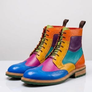 May include: A pair of ankle-high lace-up boots featuring a multi-colored leather design with blue, pink, yellow, orange, purple, and teal panels. The boots include classic brogue detailing, brown laces, a pull tab at the heel, and a green sole.