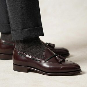 May include: A pair of dark burgundy leather tassel loafers featuring a decorative black cord lace detail across the vamp. The shoes are worn with dark grey ribbed socks and dark grey cuffed trousers, showcasing a low heel and a smooth finish.