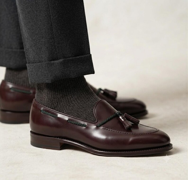 May include: A pair of dark burgundy leather tassel loafers featuring a decorative black cord lace detail across the vamp. The shoes are worn with dark grey ribbed socks and dark grey cuffed trousers, showcasing a low heel and a smooth finish.