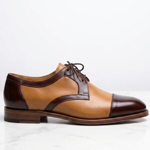 May include: A two-tone oxford shoe featuring a tan leather body with dark brown leather accents on the toe cap, heel, and lace panel. The shoe includes a matching dark brown lace-up closure and a stacked wooden heel with a dark brown finish.