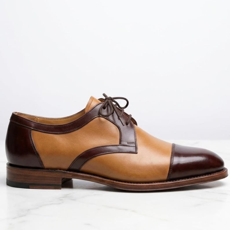 May include: A two-tone oxford shoe featuring a tan leather body with dark brown leather accents on the toe cap, heel, and lace panel. The shoe includes a matching dark brown lace-up closure and a stacked wooden heel with a dark brown finish.