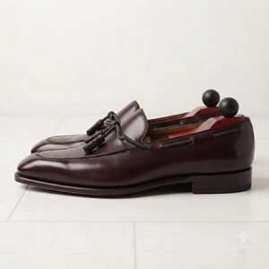 May include: A pair of dark burgundy leather loafers featuring a decorative tassel detail on the vamp, a braided leather lace accent around the collar, and a low stacked heel. Each shoe contains a wooden shoe tree with a black spherical knob.