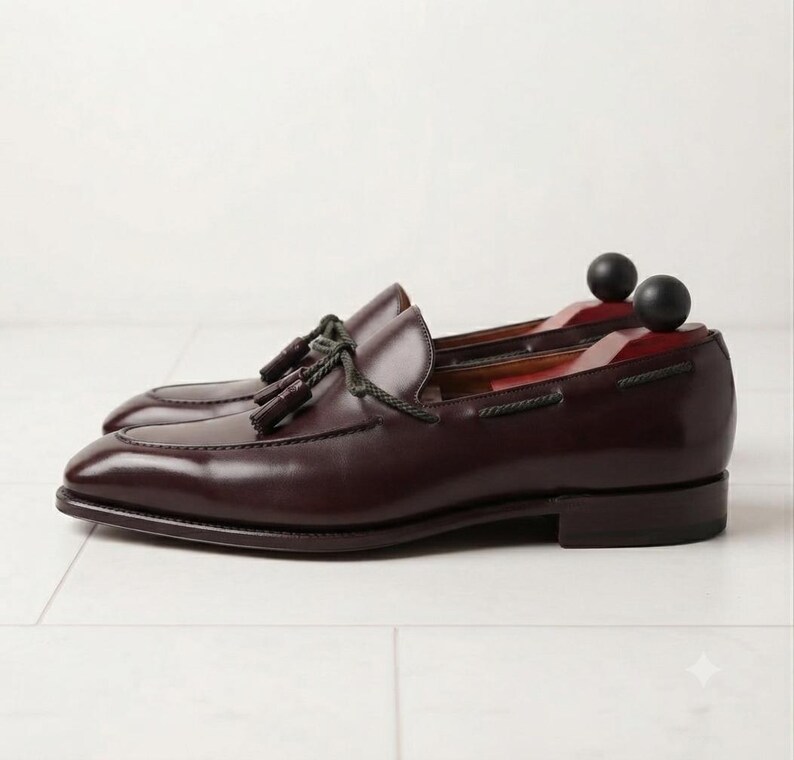 May include: A pair of dark burgundy leather loafers featuring a decorative tassel detail on the vamp, a braided leather lace accent around the collar, and a low stacked heel. Each shoe contains a wooden shoe tree with a black spherical knob.