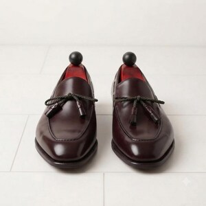 May include: A pair of dark burgundy leather loafers featuring a polished finish, a rounded toe box, and a decorative braided cord tie across the vamp with small matching tassels. Each shoe contains a red wooden shoe tree with a black spherical knob.