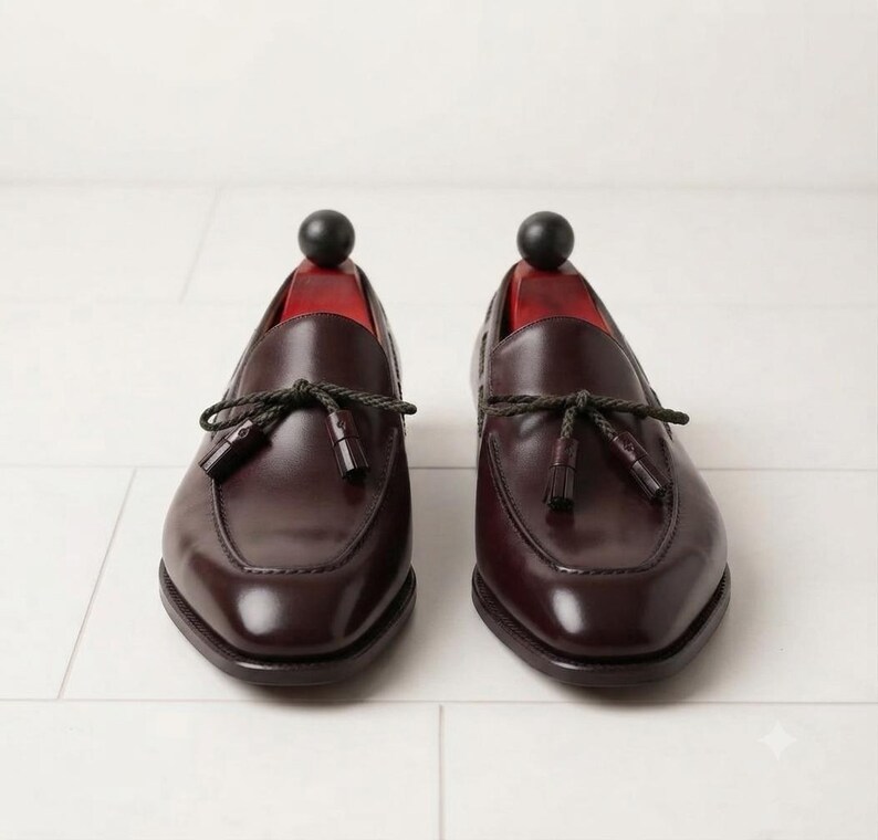 May include: A pair of dark burgundy leather loafers featuring a polished finish, a rounded toe box, and a decorative braided cord tie across the vamp with small matching tassels. Each shoe contains a red wooden shoe tree with a black spherical knob.