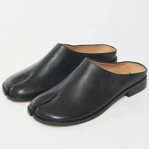 May include: A pair of black leather slip-on mules featuring a distinct split-toe design. These shoes have a smooth matte finish, a low heel, and a tan interior lining. The construction includes a central seam running from the split toe to the opening.