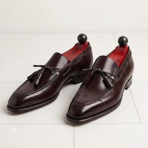 May include: A pair of dark burgundy leather tassel loafers featuring a braided leather lace detail across the vamp, a stitched apron toe, and a low stacked heel. Each shoe contains a red wooden shoe tree with a black spherical knob handle.