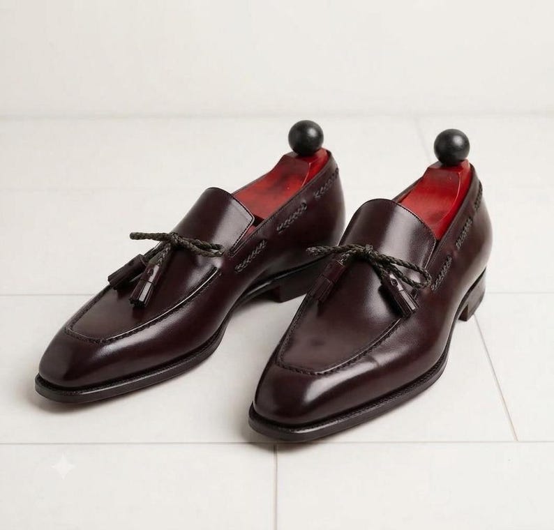May include: A pair of dark burgundy leather tassel loafers featuring a braided leather lace detail across the vamp, a stitched apron toe, and a low stacked heel. Each shoe contains a red wooden shoe tree with a black spherical knob handle.
