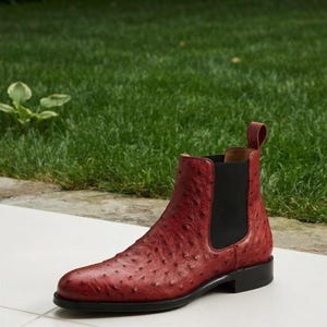May include: A red Chelsea boot featuring a textured, quill-patterned surface, a black elastic side panel, a matching red pull tab at the heel, and a solid black sole. The boot has a rounded toe and a sleek, ankle-high silhouette.