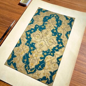 May include: A rectangular paper artwork featuring an intricate, repeating pattern of gold-colored floral and vine motifs set against a deep teal background. The design is framed by a wide, plain cream-colored border on a wooden surface.