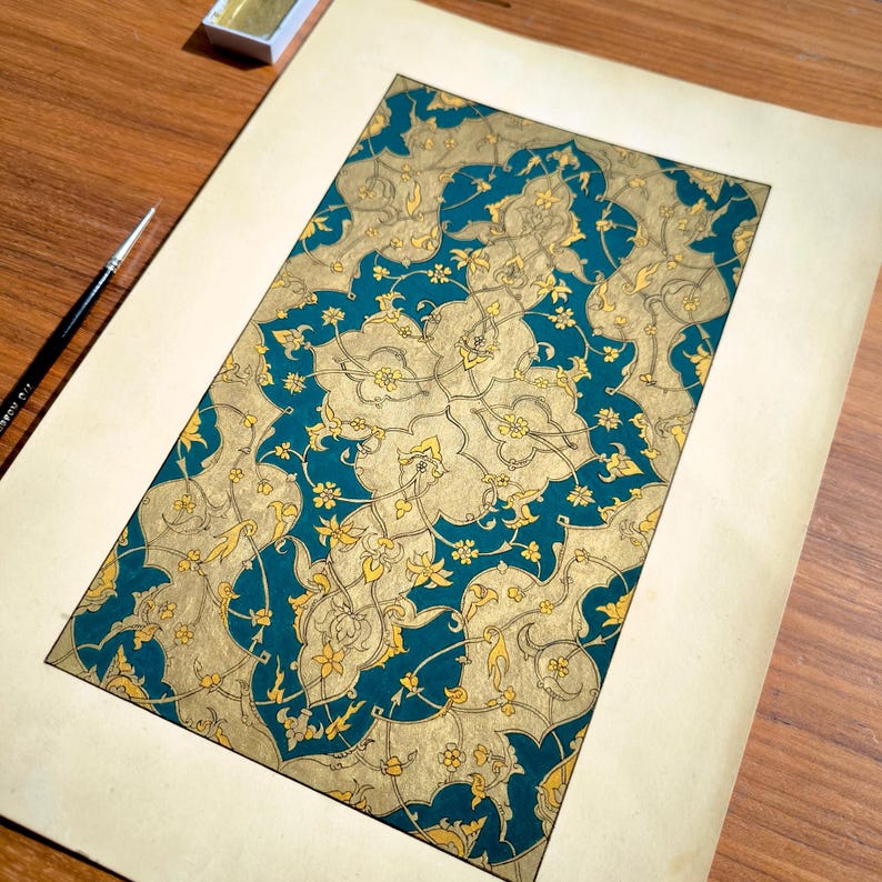 May include: A rectangular paper artwork featuring an intricate, repeating pattern of gold-colored floral and vine motifs set against a deep teal background. The design is framed by a wide, plain cream-colored border on a wooden surface.