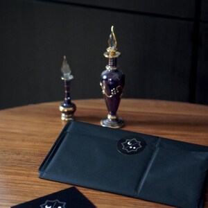 May include: A black rectangular package sealed with a circular sticker featuring a white crescent moon and two dots inside a star shape. Beside it are two purple glass perfume bottles with gold accents, a black business card, and a white card.