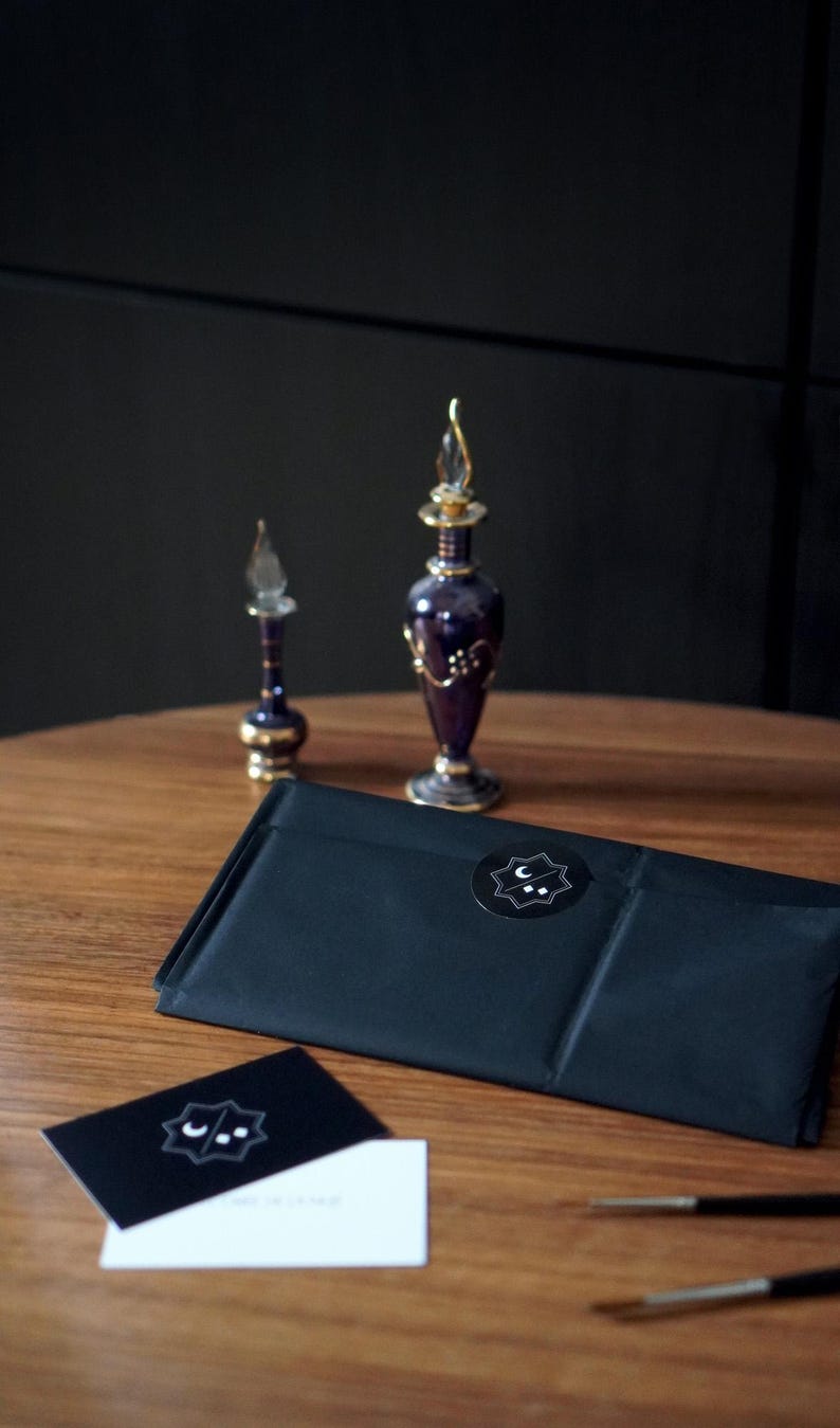 May include: A black rectangular package sealed with a circular sticker featuring a white crescent moon and two dots inside a star shape. Beside it are two purple glass perfume bottles with gold accents, a black business card, and a white card.
