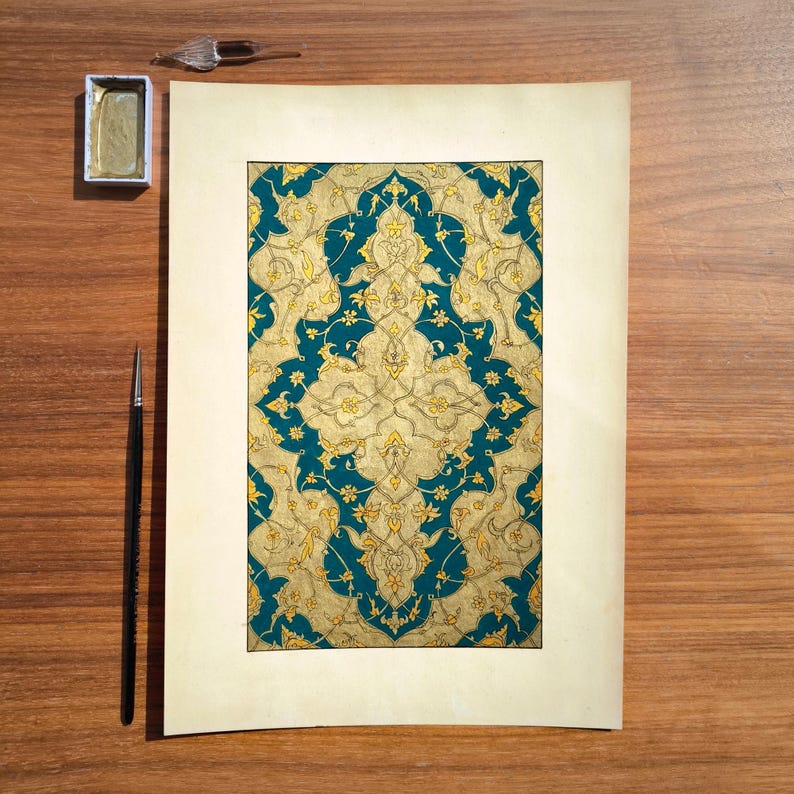May include: A rectangular paper sheet featuring an intricate, symmetrical pattern of gold floral and vine motifs set against a deep teal background. A small rectangular gold paint pan, a glass dropper, and a thin black paintbrush are positioned nearby.