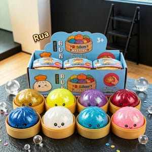 Puede incluir: Una caja expositora de bollos blanditos Rainbow Mystery con ocho juguetes de bollos coloridos y brillantes en recipientes beige individuales. Cada bollo tiene una cara sonriente. La caja muestra el texto "Rainbow Mystery", "Bun" y "Will You Find The Super Rare!"