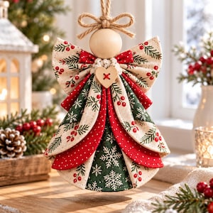 May include: A hanging angel ornament crafted from fabric with a cream background featuring red berries and green pine needles. The dress layers include red polka dot and green snowflake patterns, accented by a wooden bead head and a small heart button.