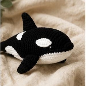 Peut inclure: Un jouet orque crocheté en noir et blanc avec une nageoire dorsale, des nageoires latérales et une tache blanche près de l'œil. La figurine en peluche est réalisée avec des points de fil visibles, a un corps arrondi et un petit œil en perle noire.