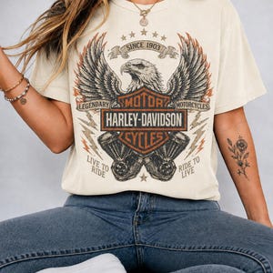 Op de afbeelding: Een cr&egrave;mekleurig T-shirt met korte mouwen en een grafische print van een Amerikaanse zeearend met gespreide vleugels boven het Harley-Davidson-logo. Het ontwerp bevat de tekst SINCE 1903, LEGENDARY MOTORCYCLES, LIVE TO RIDE en RIDE TO LIVE in oranje en zwart.