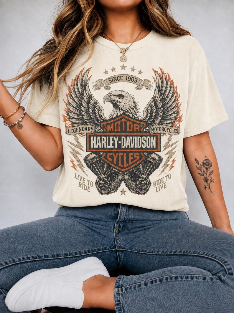 Op de afbeelding: Een cr&egrave;mekleurig T-shirt met korte mouwen en een grafische print van een Amerikaanse zeearend met gespreide vleugels boven het Harley-Davidson-logo. Het ontwerp bevat de tekst SINCE 1903, LEGENDARY MOTORCYCLES, LIVE TO RIDE en RIDE TO LIVE in oranje en zwart.
