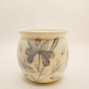 May include: A cream-coloured ceramic pot with a rounded body and a flared rim. The exterior features a hand-painted, watercolour-style floral design in shades of muted grey, depicting petals, leaves, and stems wrapping around the curved surface.