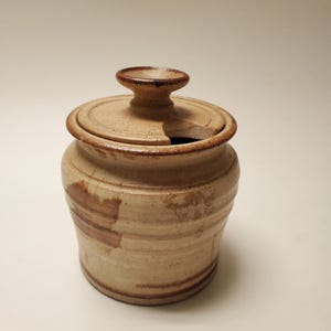 May include: A beige ceramic jar with a matching lid featuring a small knob handle and a cutout notch for a spoon. The jar has a speckled texture, horizontal ridges along the body, and darker brown glaze accents around the rim and base.