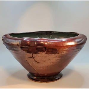 May include: A wide, deep ceramic bowl with a metallic, reddish-brown glazed exterior and a smooth, light-coloured interior. The bowl features a thick, rounded rim, a narrow base, and a distinct, dark, linear crack-like mark on the side surface.