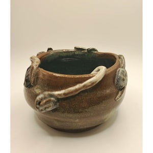 May include: A small, round, brown ceramic bowl with a dark green interior glaze. The exterior features applied, textured, light-coloured clay branch-like decorations that wrap around the rim and sides of the vessel, creating a tactile surface.