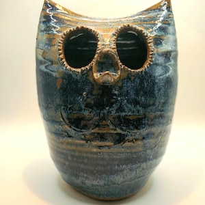 May include: A ceramic owl figurine featuring a glossy glaze in shades of deep blue, teal, and earthy brown. The piece has two large, hollow circular eye sockets, a small pointed beak, and etched wing details on the front of its rounded body.